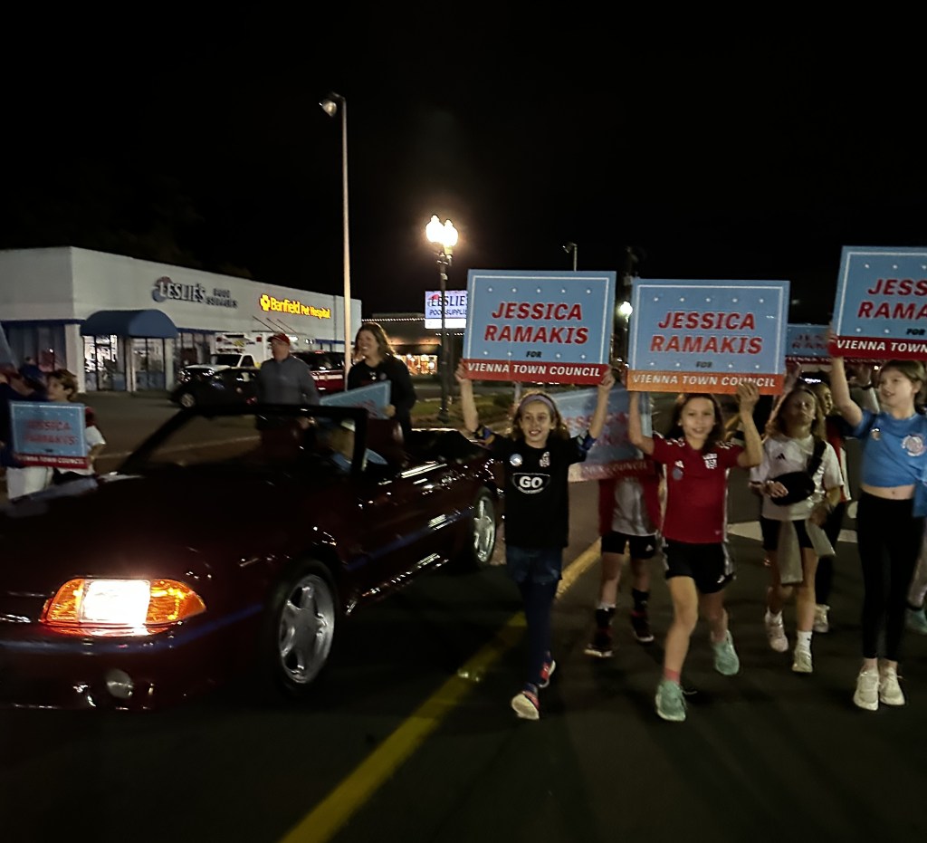 Jessica Ramakis riding in a convertible at the 2023 Vienna Halloween Parade as youth who participate in Vienna town sports hold Jessica Ramakis for Vienna Town Council Signs.